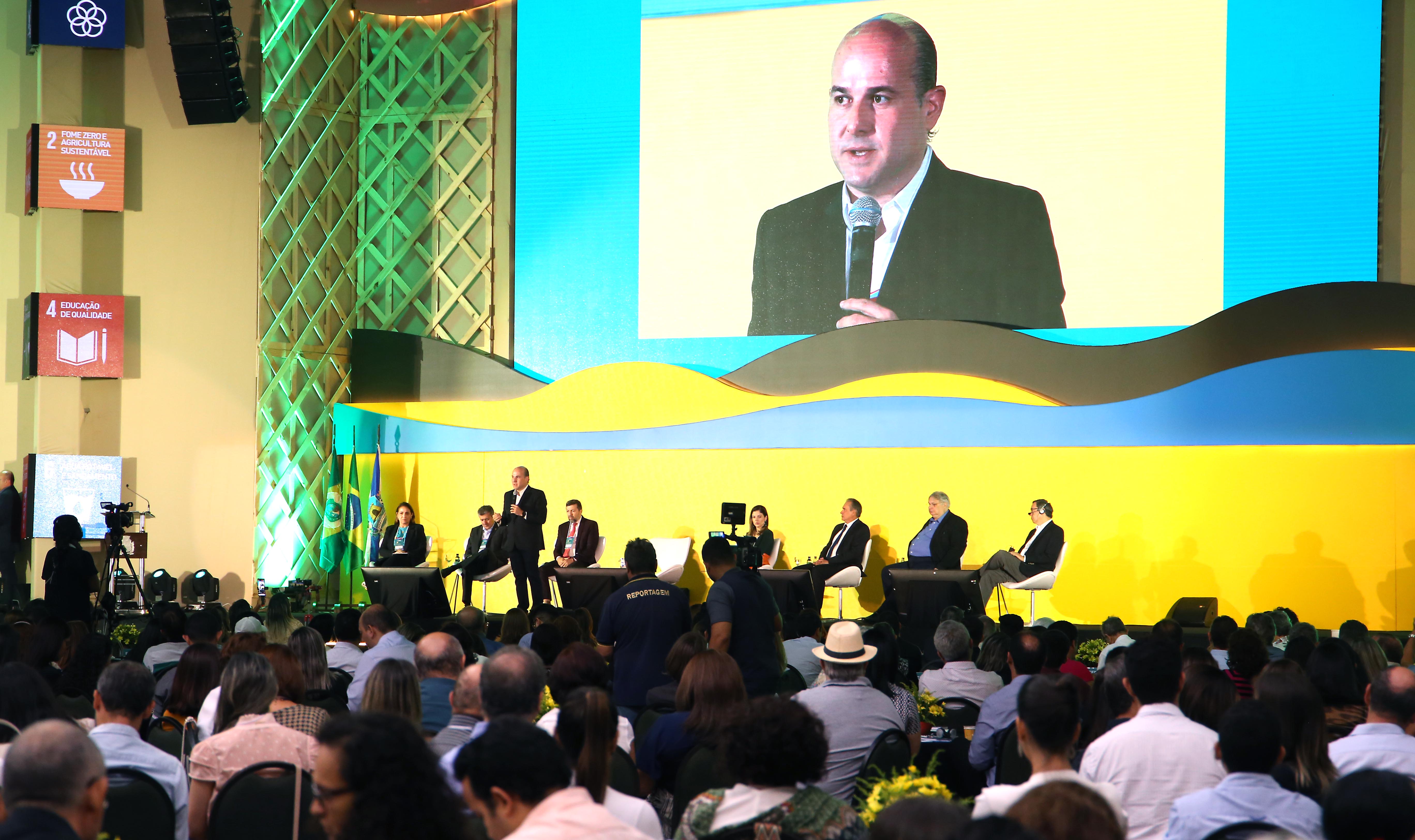 prefeito em cima de palco falando ao microfone e pessoas sentadas atrás